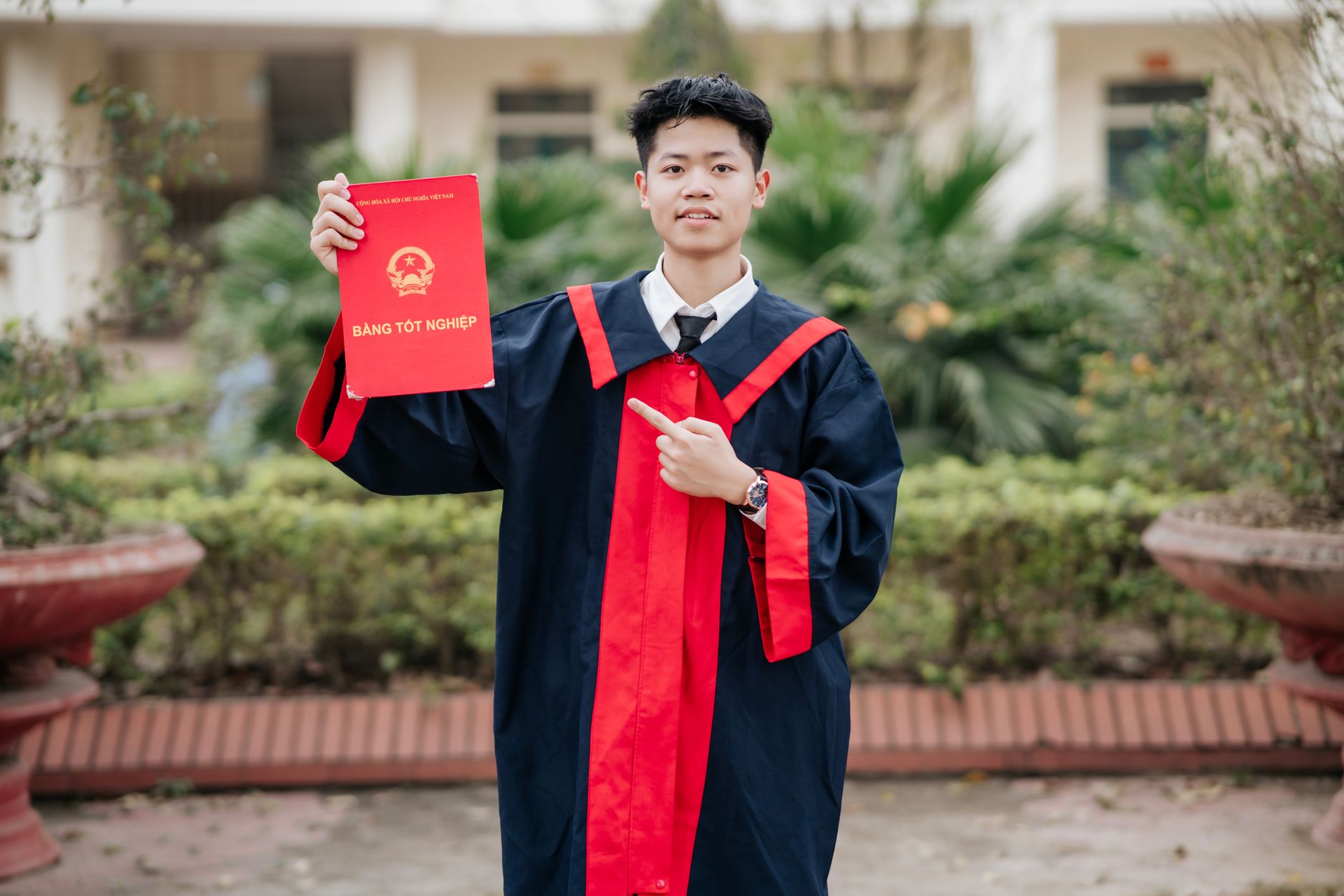 Young graduate in red-trimmed gown proudly holding diploma outdoors.