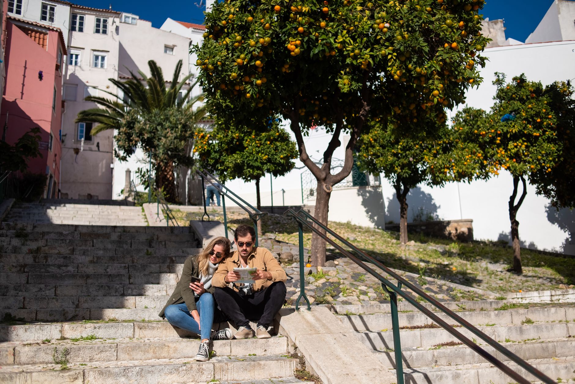 Couple sitting on steps with a map, surrounded by vibrant orange trees in a sunny urban setting.