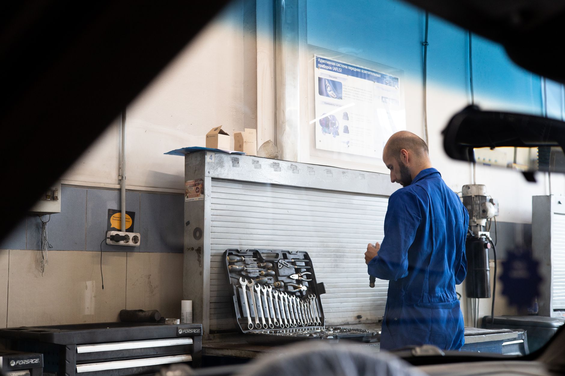 Bald mechanic in blue coveralls checking tools inside an automotive workshop.