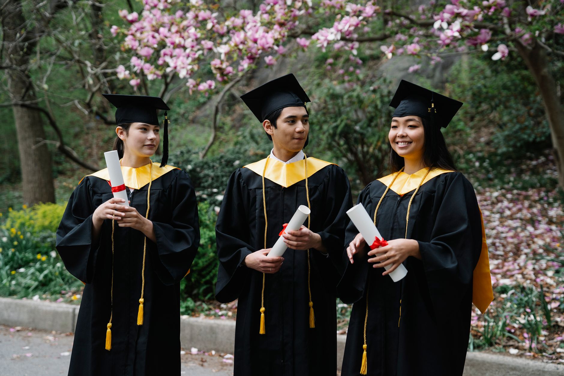 Three graduates in caps and gowns celebrating outdoors with diplomas in hand.