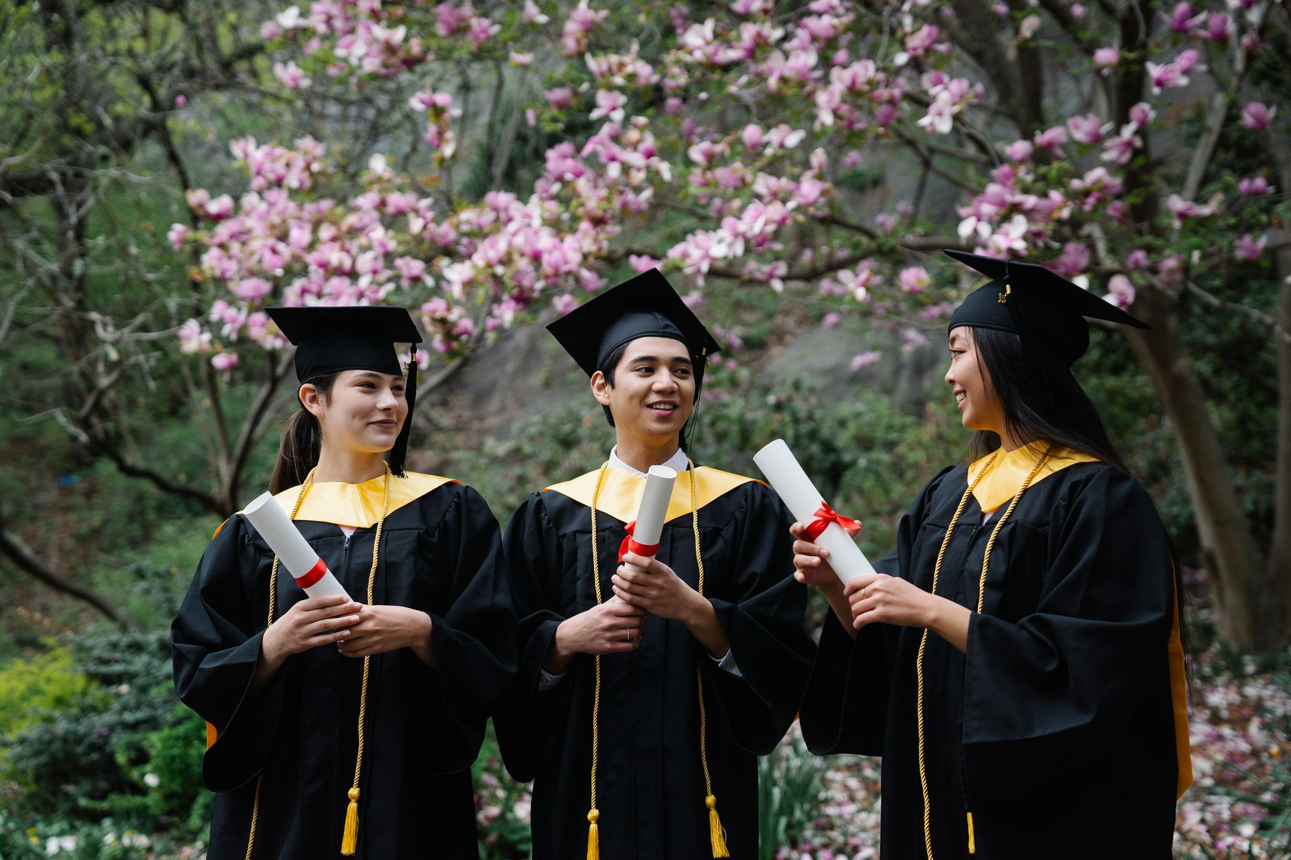 Three happy graduates celebrate outdoors in a blooming garden with diplomas and caps.