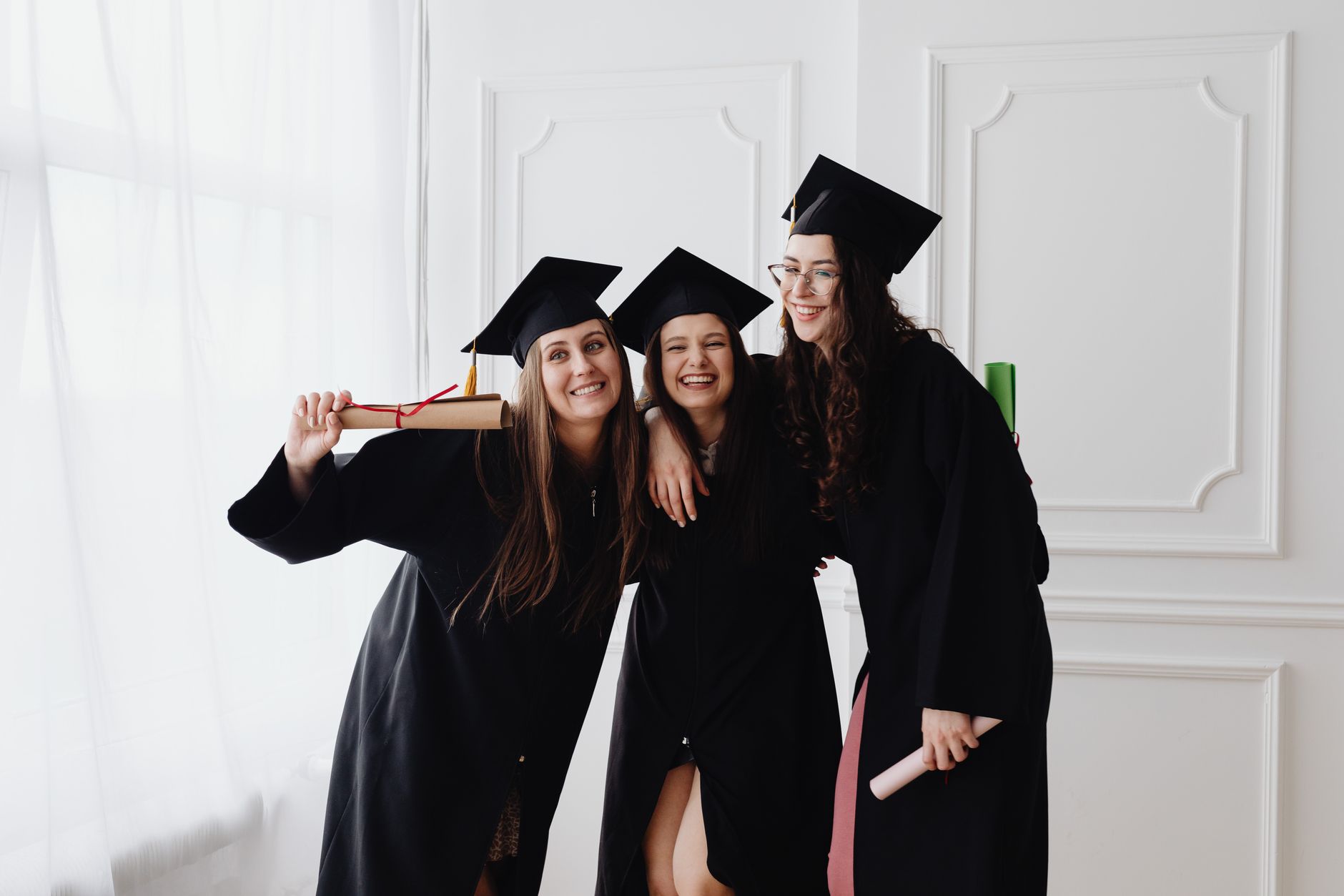 Three smiling graduates in caps and gowns celebrating indoors together.